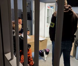 Policjant stoi za okratowanymi drzwiami, obok niego siedzi zatrzymany mężczyzna i umundurowany policjant.