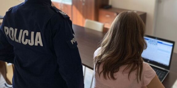 Policjant stoi przy kobiecie siedzącej przed laptopem.