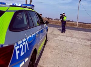 Policjant stoi przed radiowozem i mierzy prędkość przy drodze.