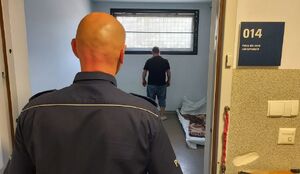 Policjant stoi w drzwiach pomieszczenia dla osób zatrzymanych, w którym przy oknie stoi zatrzymany mężczyzna.